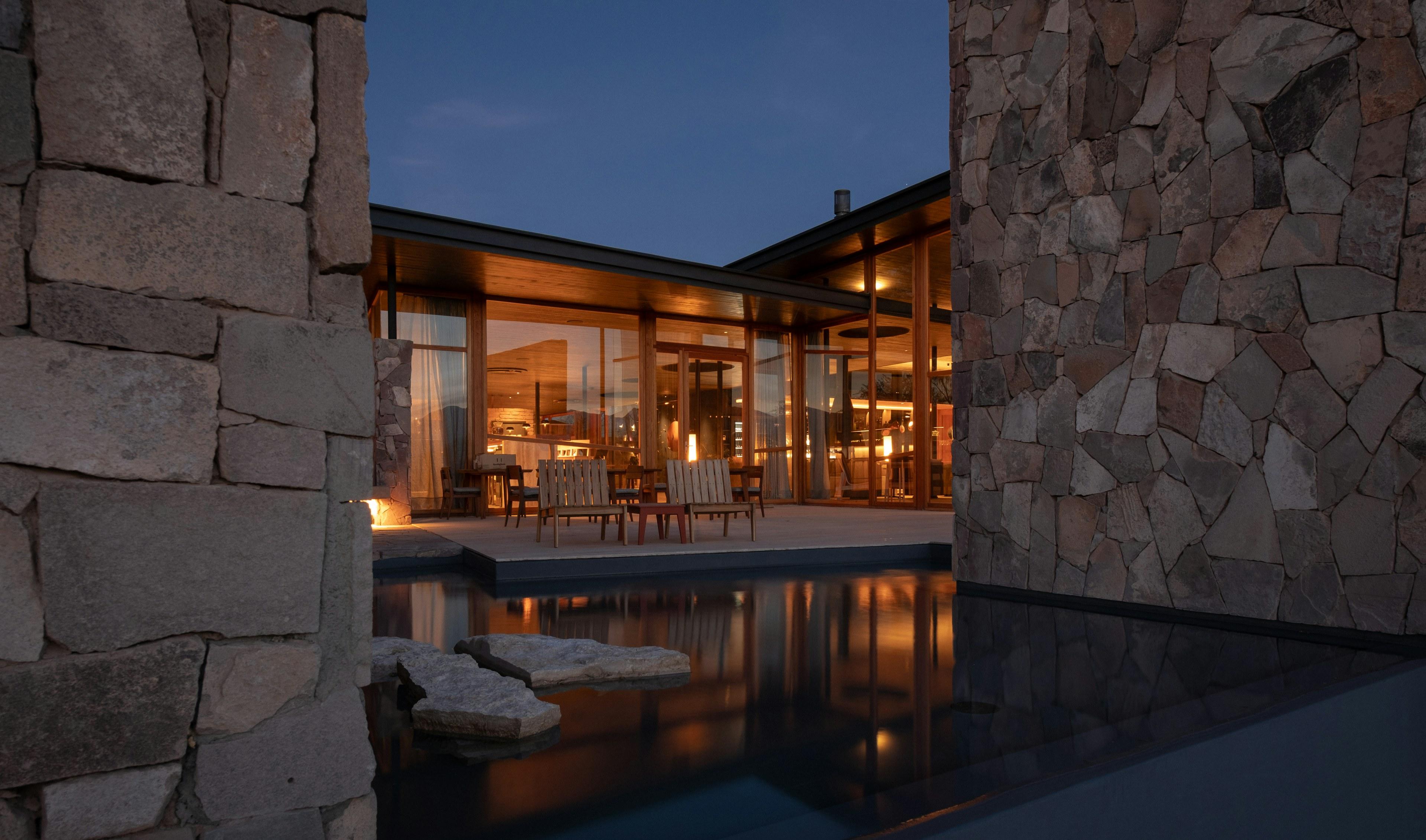 Casa contemporânea de pedra e vidro com a sala iluminada refletida na piscina ao entardecer