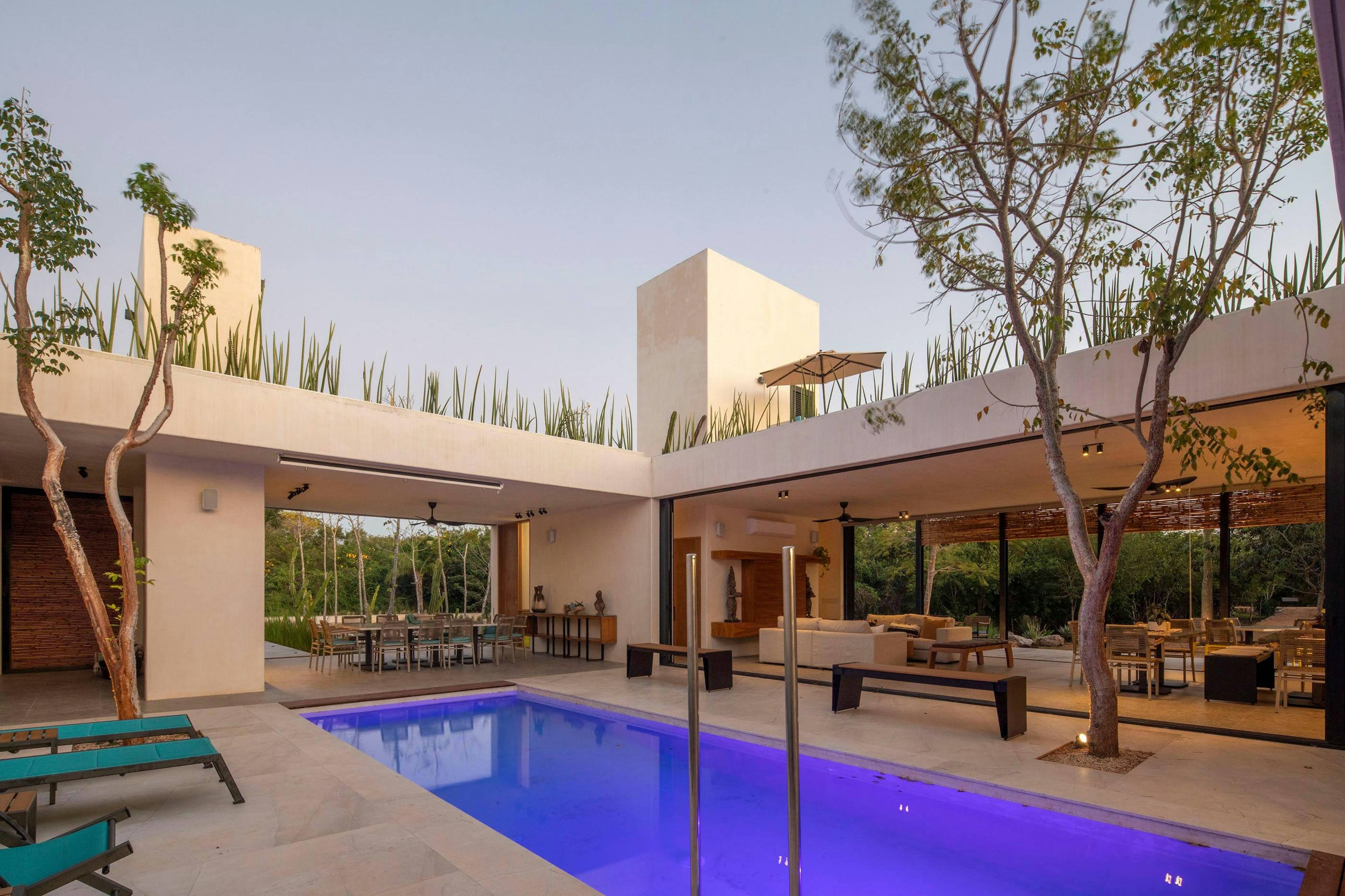 Pátio de casa moderna com piscina iluminada, árvores e varanda coberta ao entardecer